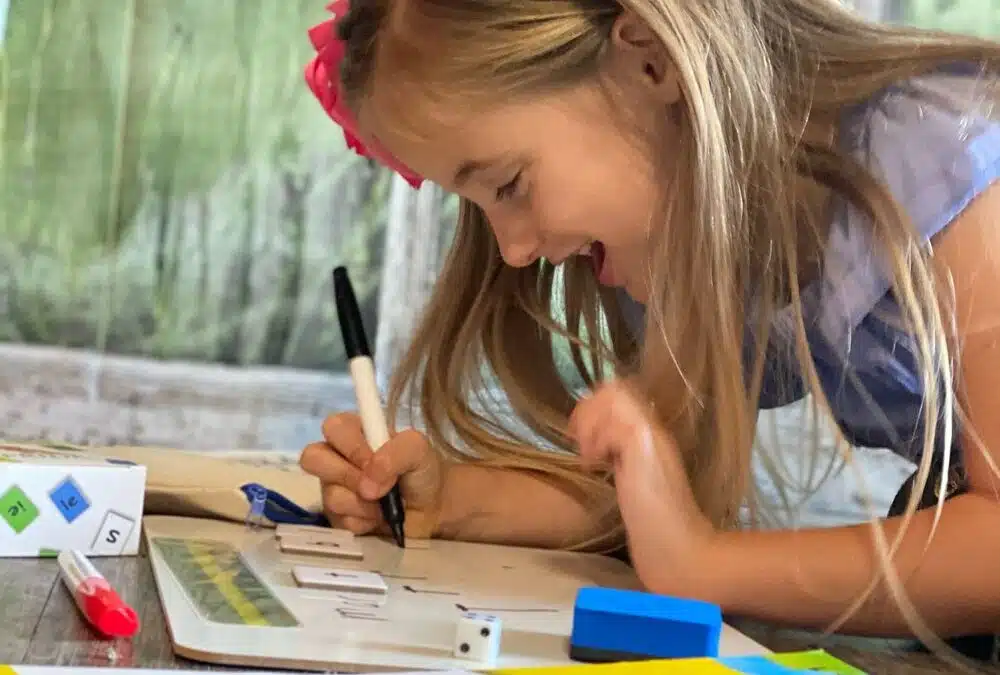 A smiling girl leans over a whiteboard to write with the PRIDE Reading Program Yellow Workbook in the foreground